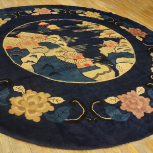 A round navy blue rug features a central landscape scene and a border of gold and rose floral motifs on wood flooring.