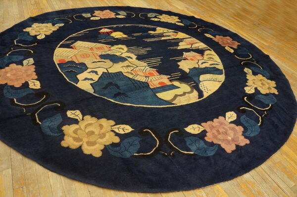 A round navy blue rug features a central landscape scene and a border of gold and rose floral motifs on wood flooring.