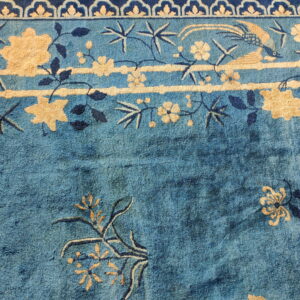 Muted blue rug with light beige and dark blue floral and avian motifs near a scalloped border.