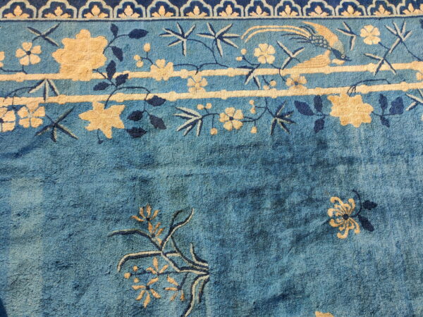 Muted blue rug with light beige and dark blue floral and avian motifs near a scalloped border.