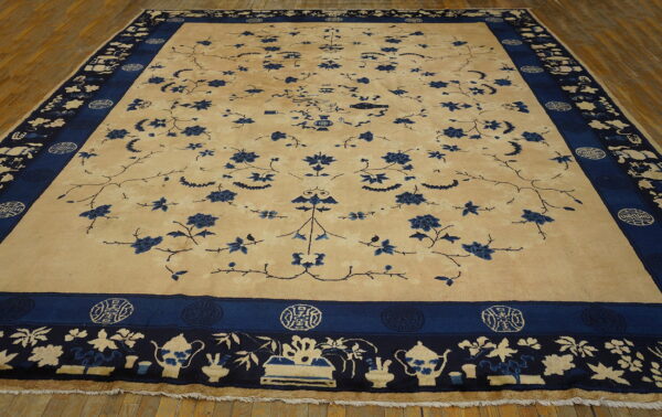 Large beige rug with sprawling blue floral motifs and complex borders featuring teapots on wood floor.