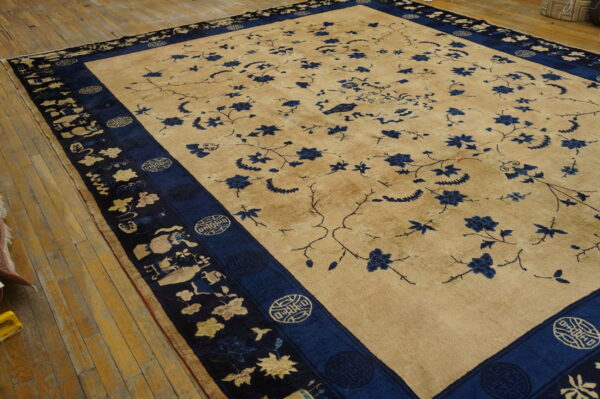 Oversize tan rug featuring scattered blue floral motifs and a complex navy pictorial border on wood flooring.