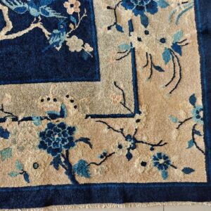 A corner of a deep indigo rug featuring a beige border and blue floral motifs on light tile flooring.