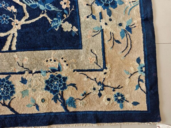 A corner of a deep indigo rug featuring a beige border and blue floral motifs on light tile flooring.