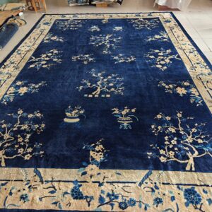Large blue rug with a beige border and flowering tree patterns, displayed on white tile floor.