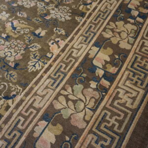 Brown rug featuring antique floral patterns in pink and cream, bordered by a beige key design, on distressed wood.