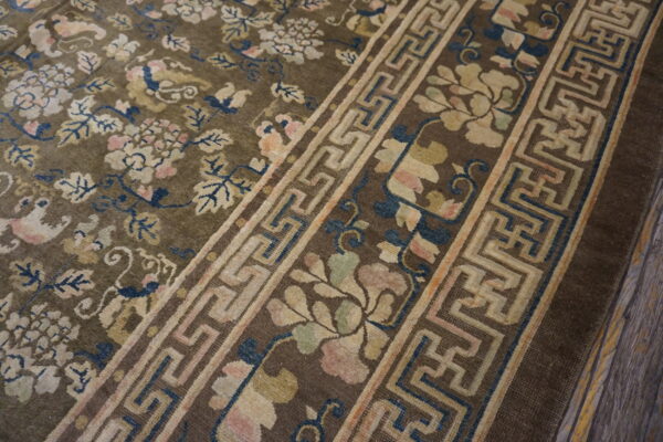 Brown rug featuring antique floral patterns in pink and cream, bordered by a beige key design, on distressed wood.