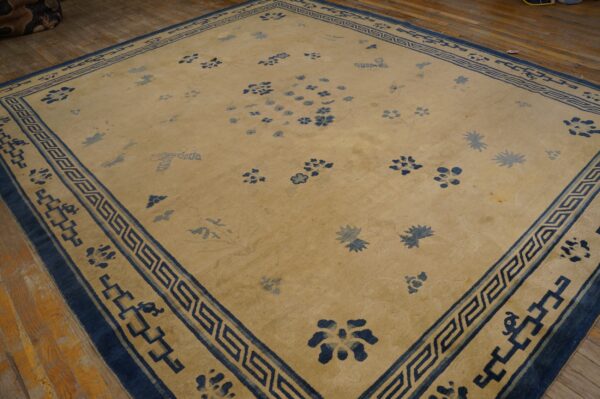 Tan rug with scattered blue floral motifs and a greek key border rests on wood planks.
