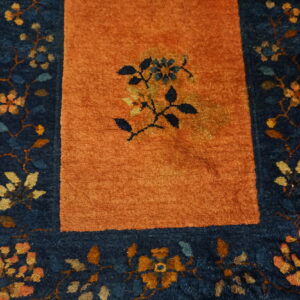 Rust colored rug featuring a central floral motif and a dark blue border on hardwood floors.