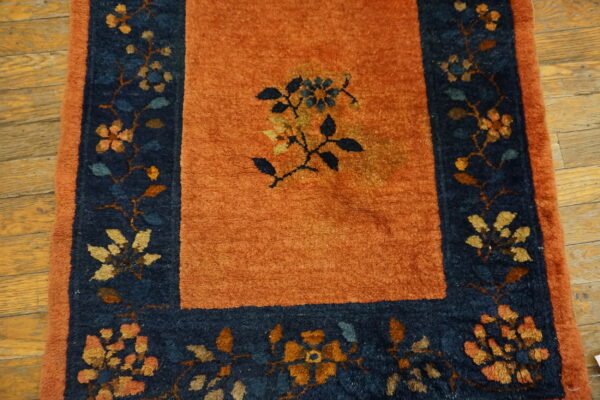Rust colored rug featuring a central floral motif and a dark blue border on hardwood floors.