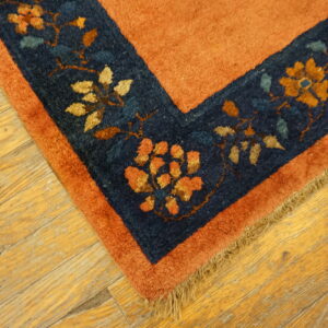 Rust rug corner with a navy blue floral border and fringe rests on light distressed wood planks.
