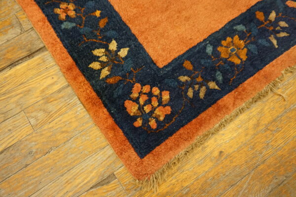 Rust rug corner with a navy blue floral border and fringe rests on light distressed wood planks.