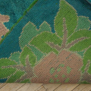 Folded rug showing thick teal, bright green, and pink floral pile resting on light wood floorboards.