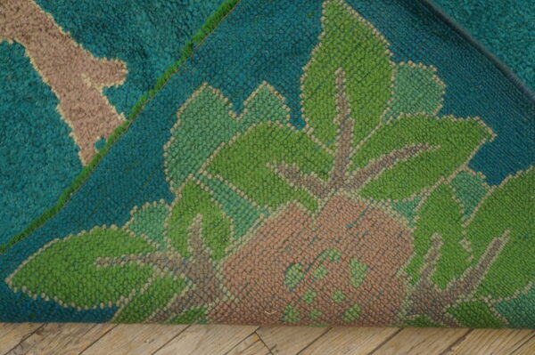 Folded rug showing thick teal, bright green, and pink floral pile resting on light wood floorboards.