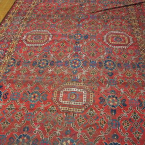 Low-pile red rug with complex blue, yellow, and brown geometric medallions on a wooden floor.