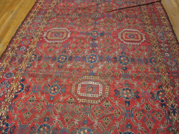 Low-pile red rug with complex blue, yellow, and brown geometric medallions on a wooden floor.