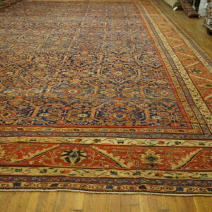 Large ornate rug featuring blue and orange colors displayed on hardwood floors next to stacked textiles.