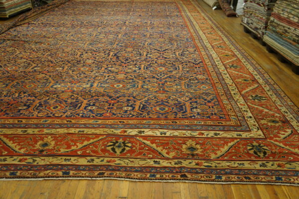Large ornate rug featuring blue and orange colors displayed on hardwood floors next to stacked textiles.