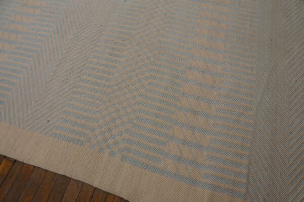 Low-pile rug in beige and light blue featuring a geometric chevron pattern on dark wood floorboards.
