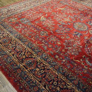 Ornate traditional rug with a deep red field and detailed blue and tan floral borders on wood flooring.