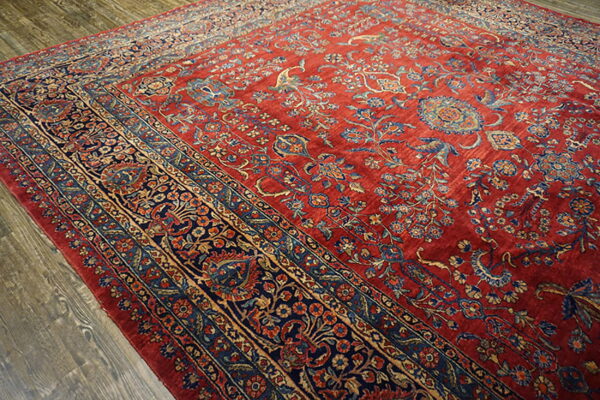 Ornate traditional rug with a deep red field and detailed blue and tan floral borders on wood flooring.