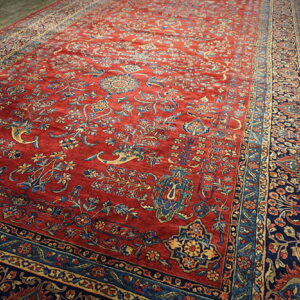 Red rug featuring intricate blue and gold floral designs and multiple borders on gray wood floors.