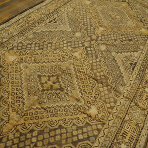 A low-pile rug with a detailed tan and dark brown geometric pattern rests on rustic wooden flooring.