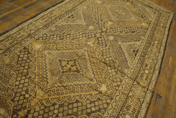 A low-pile rug with a detailed tan and dark brown geometric pattern rests on rustic wooden flooring.