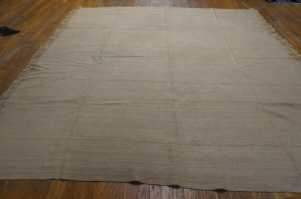 A large light beige flatweave rug with fringes and horizontal stitched seams lies on a hardwood floor.