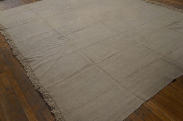 Monochromatic light beige flatweave rug with stitched seams and fringe rests on a dark brown hardwood floor.