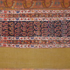A colorful, patterned rug section with navy and red geometric motifs is displayed on an olive green textile.