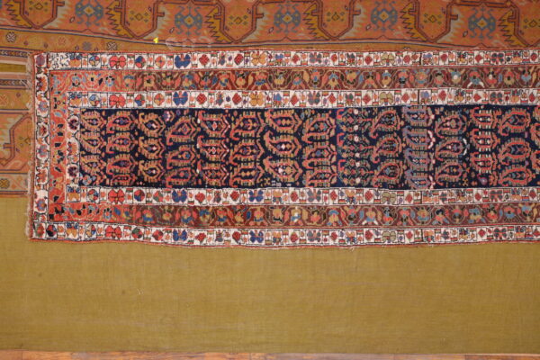 A colorful, patterned rug section with navy and red geometric motifs is displayed on an olive green textile.