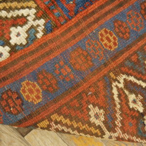 High-pile red, blue, and cream rug folded back to show a tight flat-woven border over rustic wood floor.