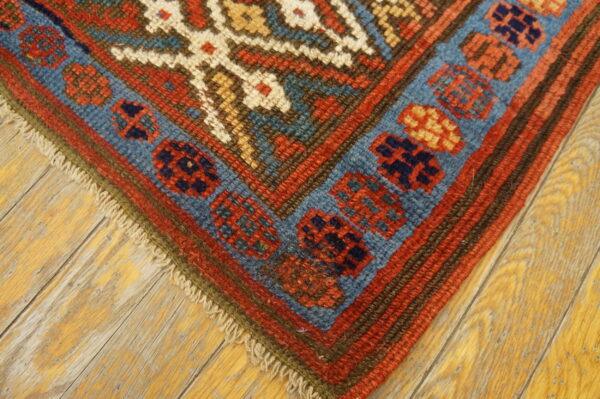 Geometric red and blue border design on a low pile rug resting on distressed hardwood planks.