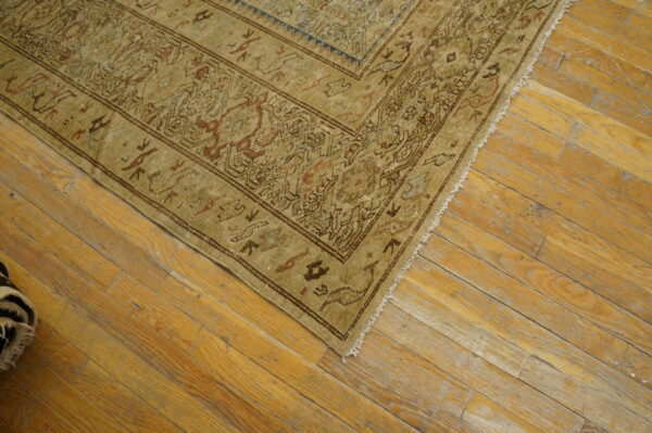 Corner of a muted tan rug with traditional brown and red patterns on distressed wood flooring.