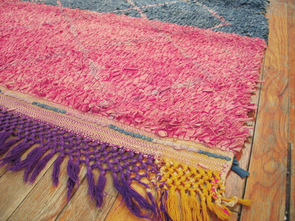 Textured rag rug in bright pink and navy, featuring purple and yellow macrame fringe on wood flooring.