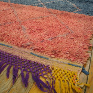 Close-up of a colorful pink and blue textured rag rug with thick purple and yellow knotted fringe on a wood floor.