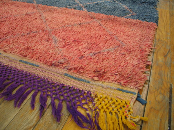 Close-up of a colorful pink and blue textured rag rug with thick purple and yellow knotted fringe on a wood floor.
