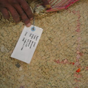 High-texture woven rug in muted beige shows colorful fabric scraps and a hand holding an attached tag.