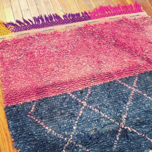 Pink and blue textured shag rug with multicolored fringe rests on natural wood plank flooring.