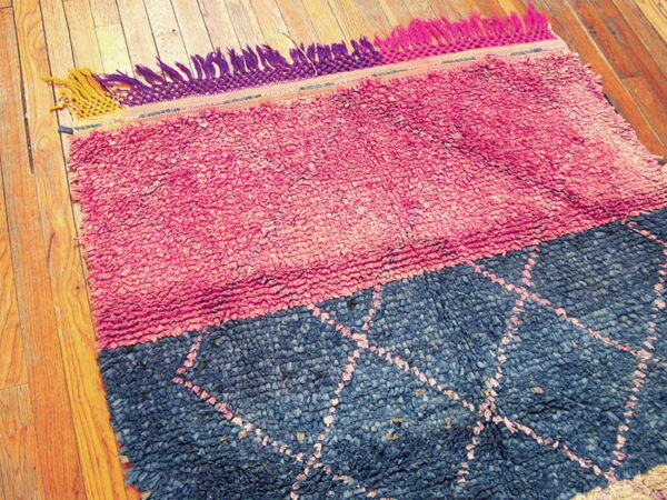Pink and blue textured shag rug with multicolored fringe rests on natural wood plank flooring.