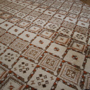 Off-white shaggy rug with brown and orange geometric diamond motifs rests on light brown wood flooring.