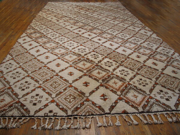 Cream high-pile rug with geometric brown and orange diamond patterns, featuring braided tassels on wood flooring.