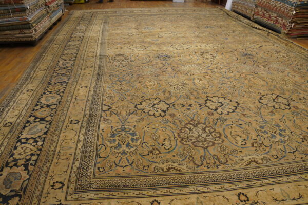 Oversize, densely patterned tan rug displayed on hardwood flooring surrounded by tall stacks of folded rugs.