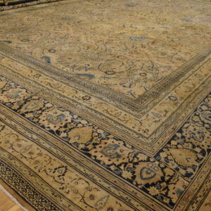 Tan and gold traditional rug featuring intricate blue and black botanical patterns, displayed on wood plank flooring.