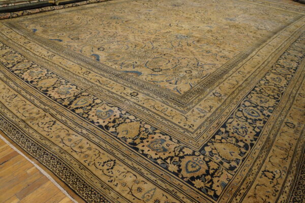 Tan and gold traditional rug featuring intricate blue and black botanical patterns, displayed on wood plank flooring.