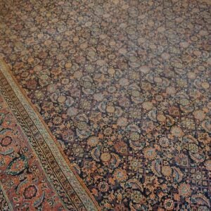A dense, low-pile rug displays a dark floral field and a wide, patterned rust-red border on wood.