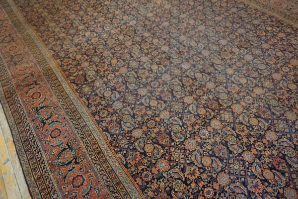 A dense, low-pile rug displays a dark floral field and a wide, patterned rust-red border on wood.