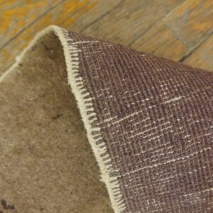 A brown rug is curled up revealing its dark woven backing on worn yellow painted wood planks.