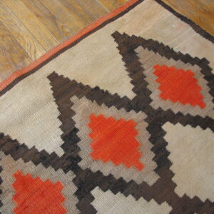 Beige flatweave rug with orange and brown stepped patterns lying diagonally on light wood flooring.
